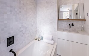 Une salle de bain moderne avec un mur recouvert de carreaux blancs, une grande baignoire, un miroir et une étagère en bois au-dessus du lavabo.