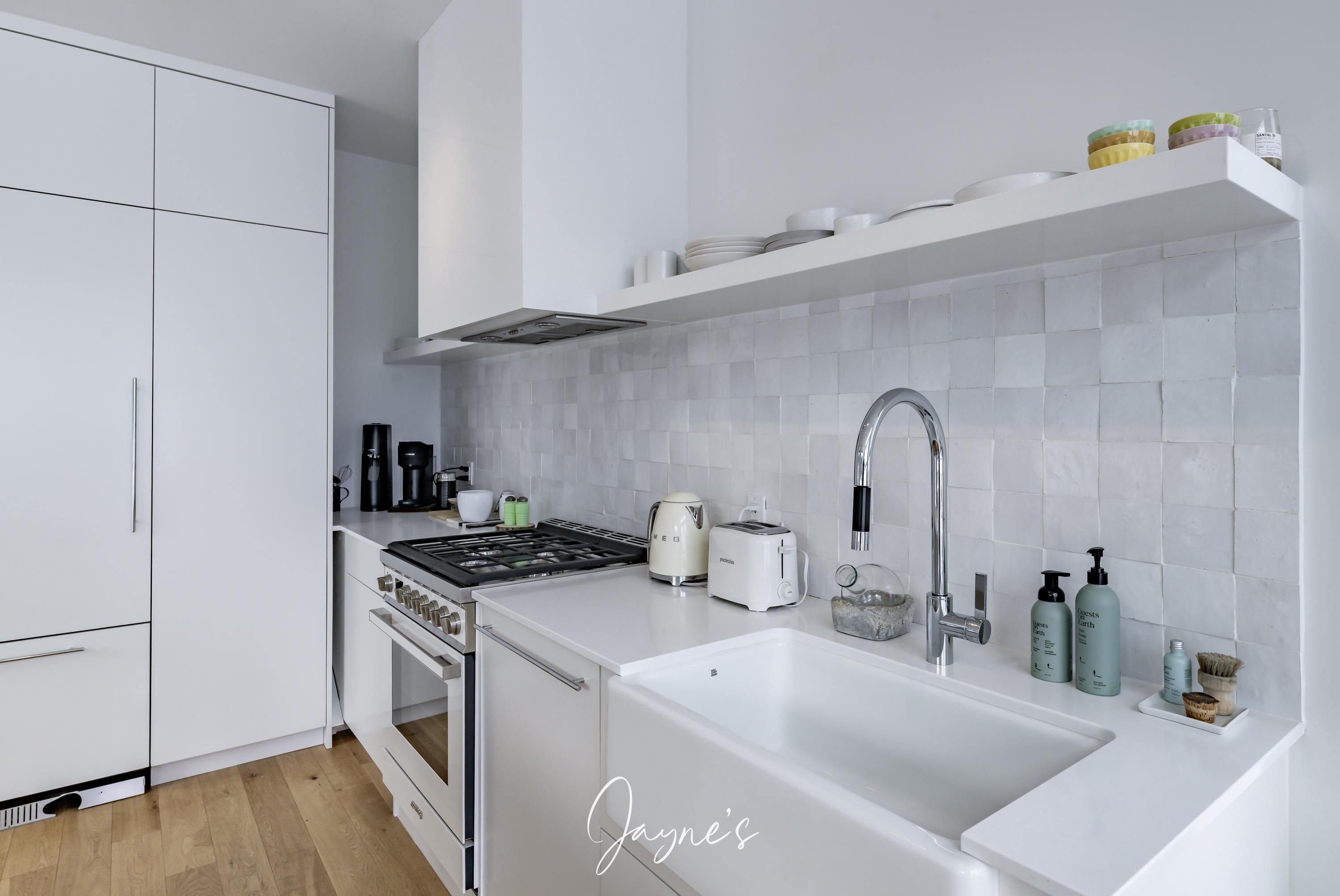 Une cuisine moderne avec des meubles blancs, une hotte en acier inoxydable, un évier et un plan de travail agrémenté de divers appareils et de bocaux de rangement.
