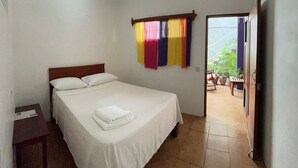 Une chambre à coucher comprenant un lit, un ventilateur de plafond, une fenêtre avec des rideaux colorés et une porte donnant sur un espace extérieur agrémenté de plantes.