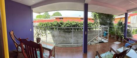 Un balcón techado con suelo de baldosas, dos sillas de madera, una mesita y plantas en macetas.