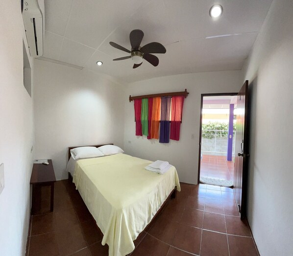 A bedroom with a bed, a ceiling fan, a window with colorful curtains, and a door leading outside. - Casa Romano Hostal (San Juan del Sur)