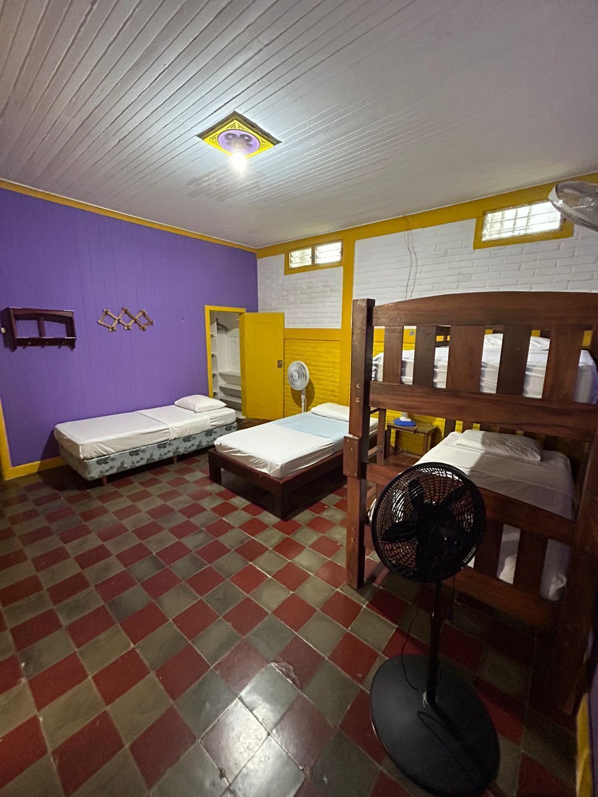 Quarto em dormitório com duas camas de solteiro, uma beliche, um ventilador e uma janela com molduras amarelas.