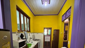 Une cuisine étroite aux murs jaunes, avec un plafond blanc et une fenêtre. On y trouve un four à micro-ondes, un évier et une petite table recouverte d’une nappe blanche.