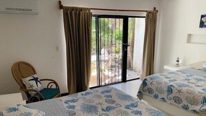 A hotel room with two beds, a wicker chair, a small table, and a sliding glass door leading to an outdoor area.