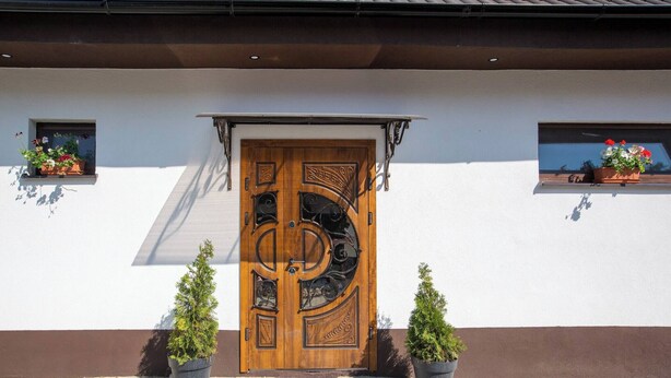 Eine Holztür mit aufwendigen Schnitzereien, begleitet von Topfpflanzen, und ein Fenster mit Blumenkasten an der Gebäudefassade.