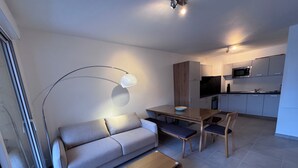 A modern living room with a sofa, coffee table, and dining table. There is a kitchen area with cabinets and a microwave. - New apartment near Prorel (Briançon)