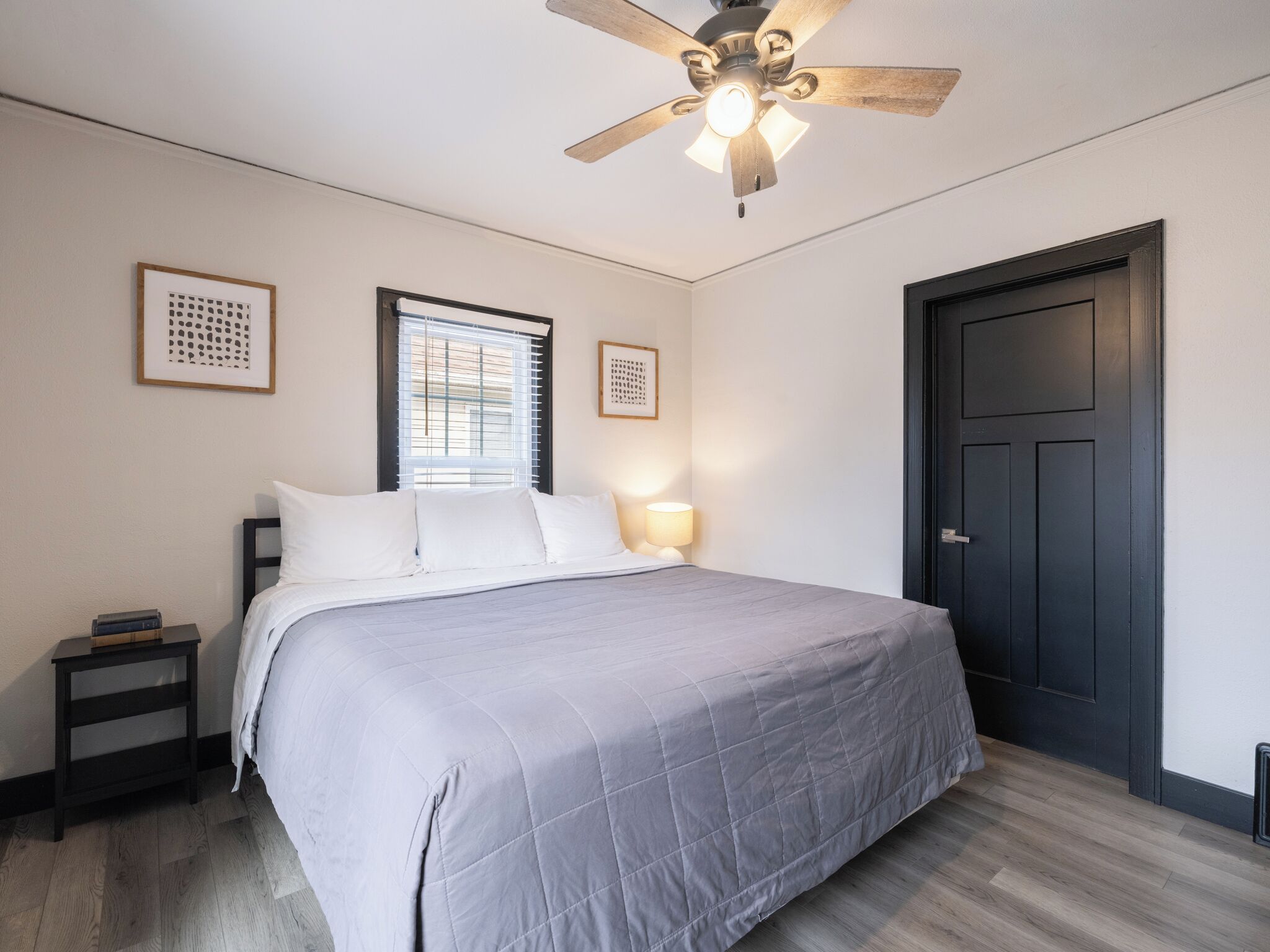 A bedroom with a bed, a nightstand, a ceiling fan, a window with blinds, and a door.