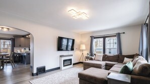 Una sala de estar moderna con chimenea, televisor montado en la pared y un comedor visible a través de un arco.