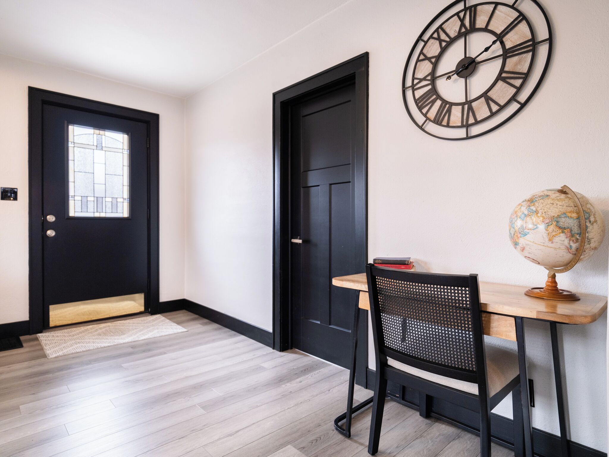 A hallway with a black door, a wooden floor, a black chair, a wooden table, and a wall clock.