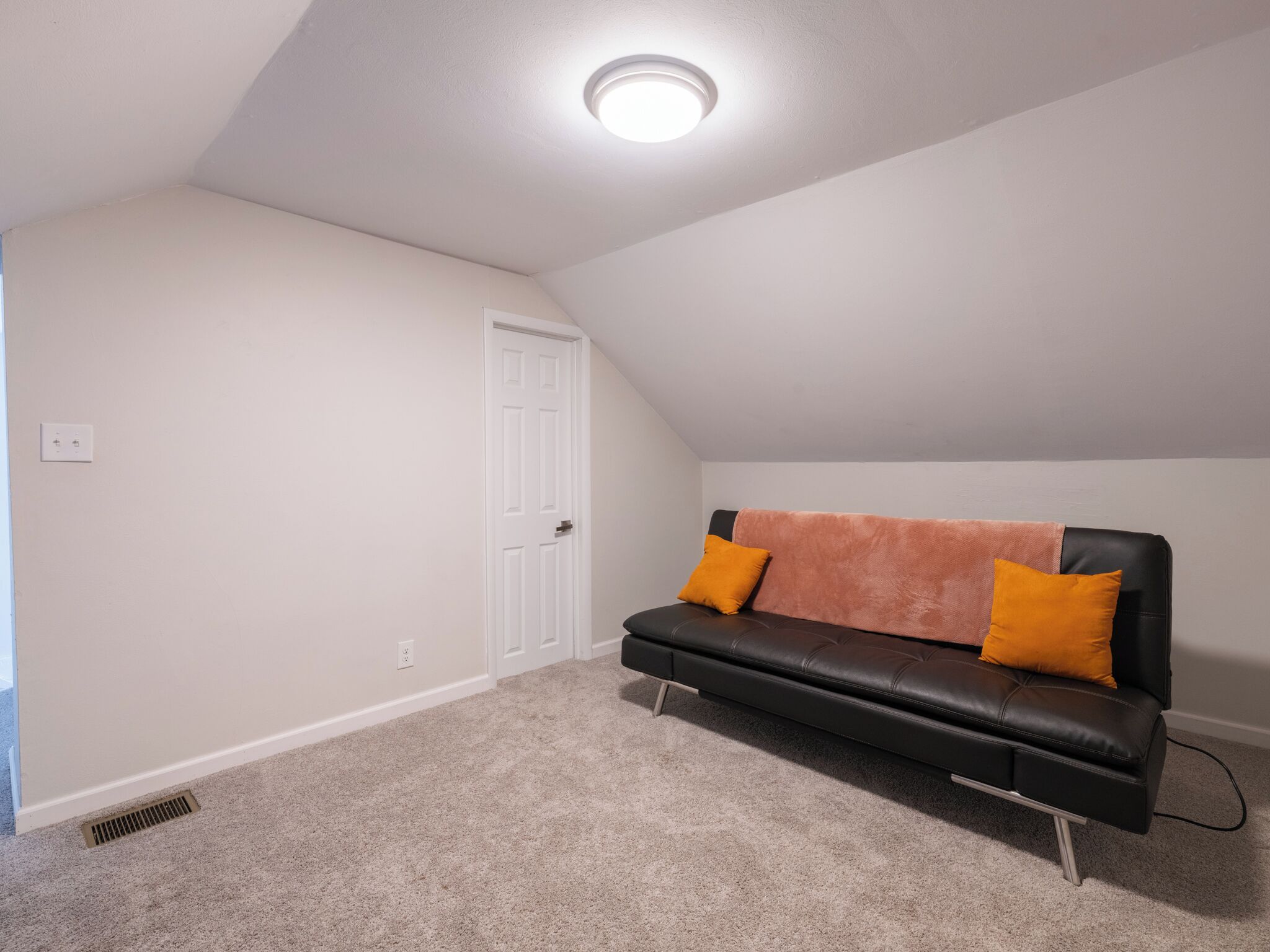 Une pièce avec un canapé en cuir noir, deux coussins orange, une porte blanche et un luminaire au plafond.