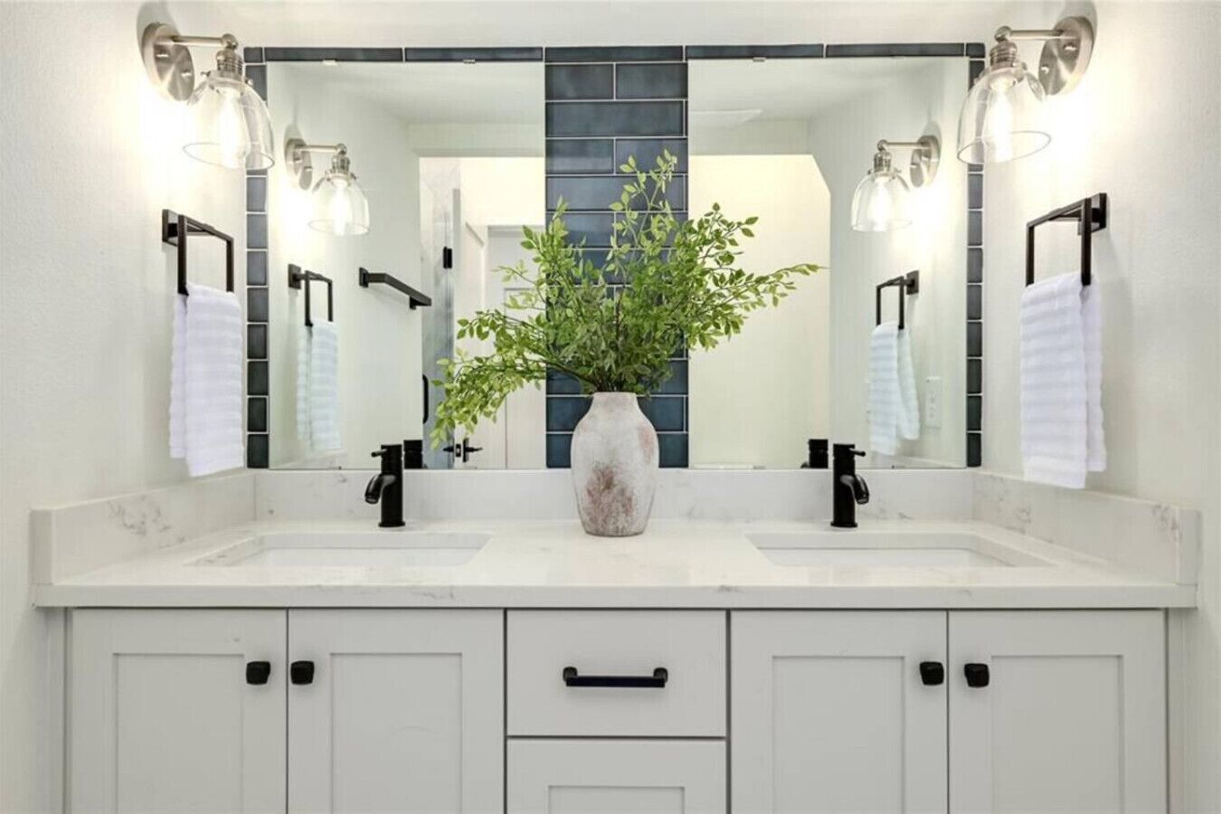 Une salle de bain avec un double vasque, un grand miroir et un vase contenant des plantes vertes.