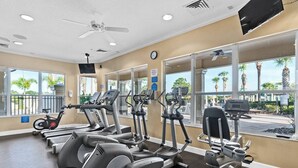 Et fitnessrum med forskellige træningsmaskiner, en ventilator i loftet og store vinduer med udsigt til palmer og en parkeringsplads.