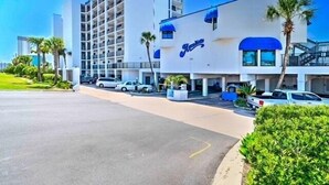Un hotel con fachada blanca, toldos azules y un estacionamiento con varios coches.