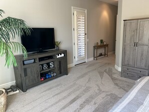 Une pièce avec un téléviseur posé sur un meuble en bois, une porte avec des stores, une petite table avec une plante et une armoire.