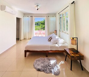 A bedroom with a bed, a bedside table, a lamp, and a view of a balcony with a table and chairs. - Spacious Family Home Near Beaches, Surf, Restaurants & Nature (rincón)