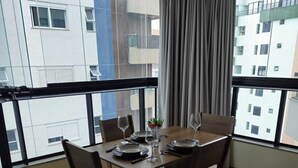 Un comedor con una mesa dispuesta para cuatro personas, ventanas amplias con cortinas y vistas a otros edificios.