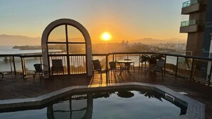 Una piscina en la azotea con vistas al atardecer, rodeada de sillones de descanso y una barandilla de cristal.