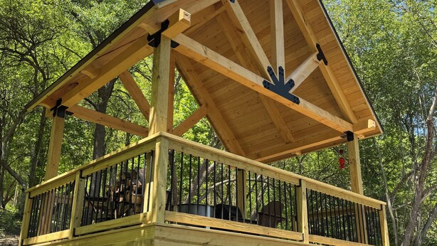 A wooden gazebo with a stone base, surrounded by trees.