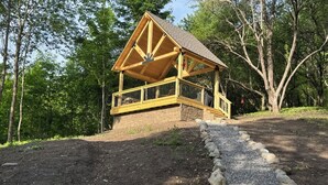 A wooden cabin with a balcony, surrounded by trees and a gravel path.