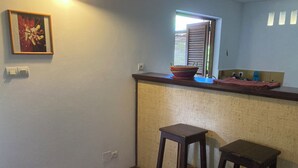 A small kitchen with a wooden countertop, two stools, a window with blinds, and a framed picture on the wall.