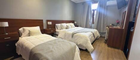 Habitación de hotel con dos camas, cabecero de madera, televisor de pantalla plana, escritorio con lámpara y ventana con cortinas.