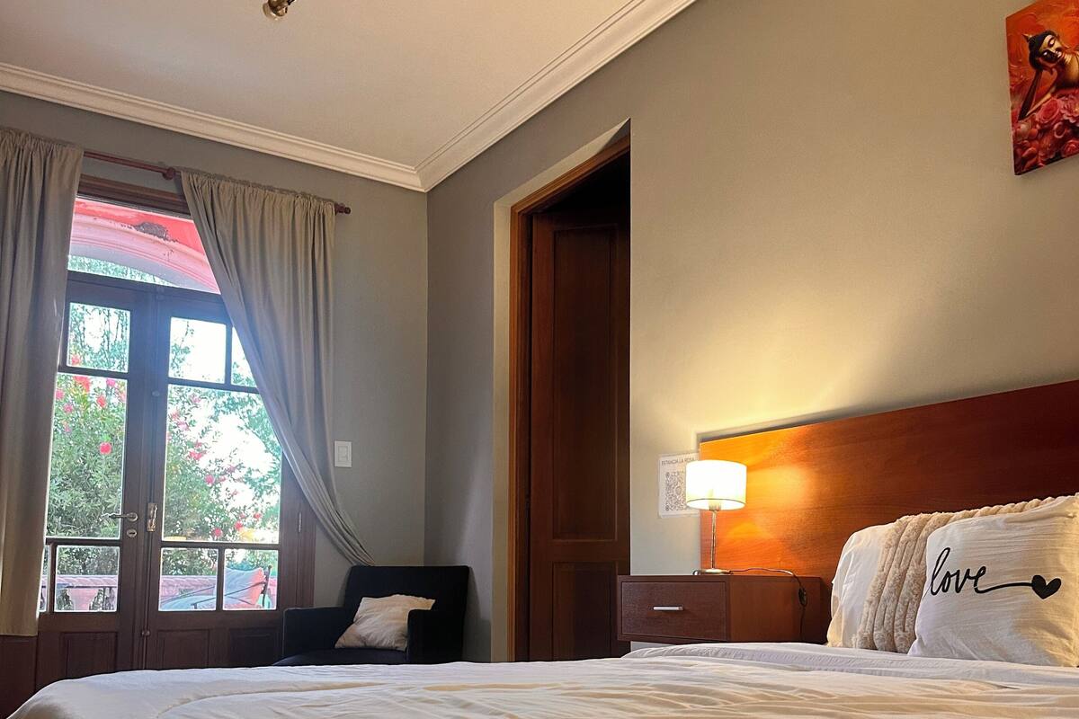 A hotel room with a large bed, a nightstand, a lamp, a window with curtains, and a door.