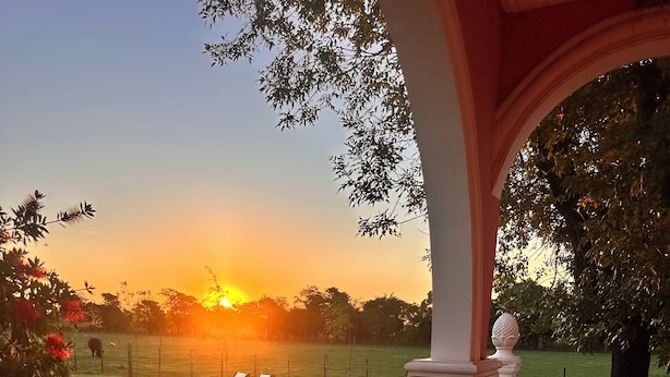 A covered outdoor seating area with a table and chairs, overlooking a grassy field and a sunset. - ESTANCIA LA ROSADITA (San Andres de Giles)