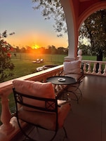 Área de estar coberta ao ar livre, com mesa e cadeiras, com vista para um campo gramado e um pôr do sol.