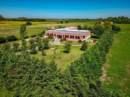 A large, single-story house with a flat roof, surrounded by dense trees and open fields.