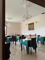 Uma sala de jantar com mesas dispostas para seis pessoas, cada uma com toalha de mesa verde e cadeiras pretas. Dois lustres pendem do teto.
