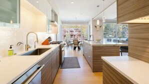 Une cuisine moderne dotée d’un îlot central, d’un coin repas avec des chaises et d’une vue sur les maisons grâce à de grandes fenêtres.