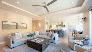 Un salon spacieux avec un grand canapé, une table basse et un ventilateur de plafond. Au fond, on aperçoit un coin cuisine avec une table à manger et des chaises.
