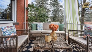 A covered outdoor seating area with wicker furniture, a checkered floor, and a view of trees and a building in the background. - Historic Gem | Soaking Tub • Fire Pit • EV Charger (Pittsburgh)