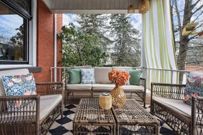 A covered outdoor seating area with wicker furniture, a checkered floor, and a view of trees and a building in the background. - Historic Gem | Soaking Tub • Fire Pit • EV Charger (Pittsburgh)