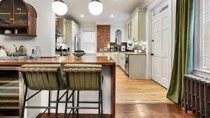 A modern kitchen with a central island, bar stools, pendant lighting, and a brick accent wall. - Historic Gem | Soaking Tub • Fire Pit • EV Charger (Pittsburgh)