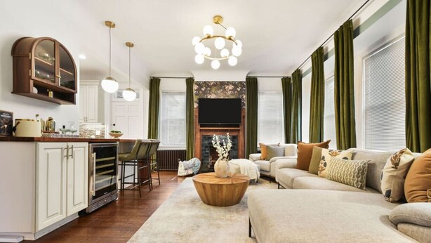 A spacious living room with a large sofa, a coffee table, and a fireplace. There is a kitchen area with a bar and chairs. - Historic Gem | Soaking Tub • Fire Pit • EV Charger (Pittsburgh)