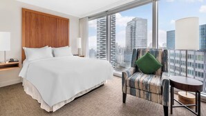 Quarto de hotel com uma cama grande, uma cadeira, uma mesinha e vista para prédios altos.