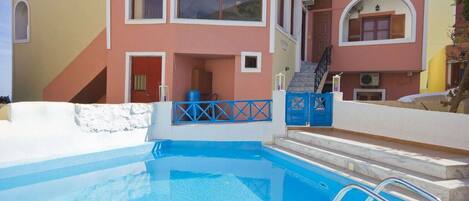 A swimming pool with a blue deck, a white wall, and a pink building with a staircase and a blue gate.