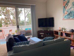 Un salon comprenant un canapé, un téléviseur, une bibliothèque et une porte-fenêtre coulissante donnant sur un espace extérieur avec une table et des chaises.
