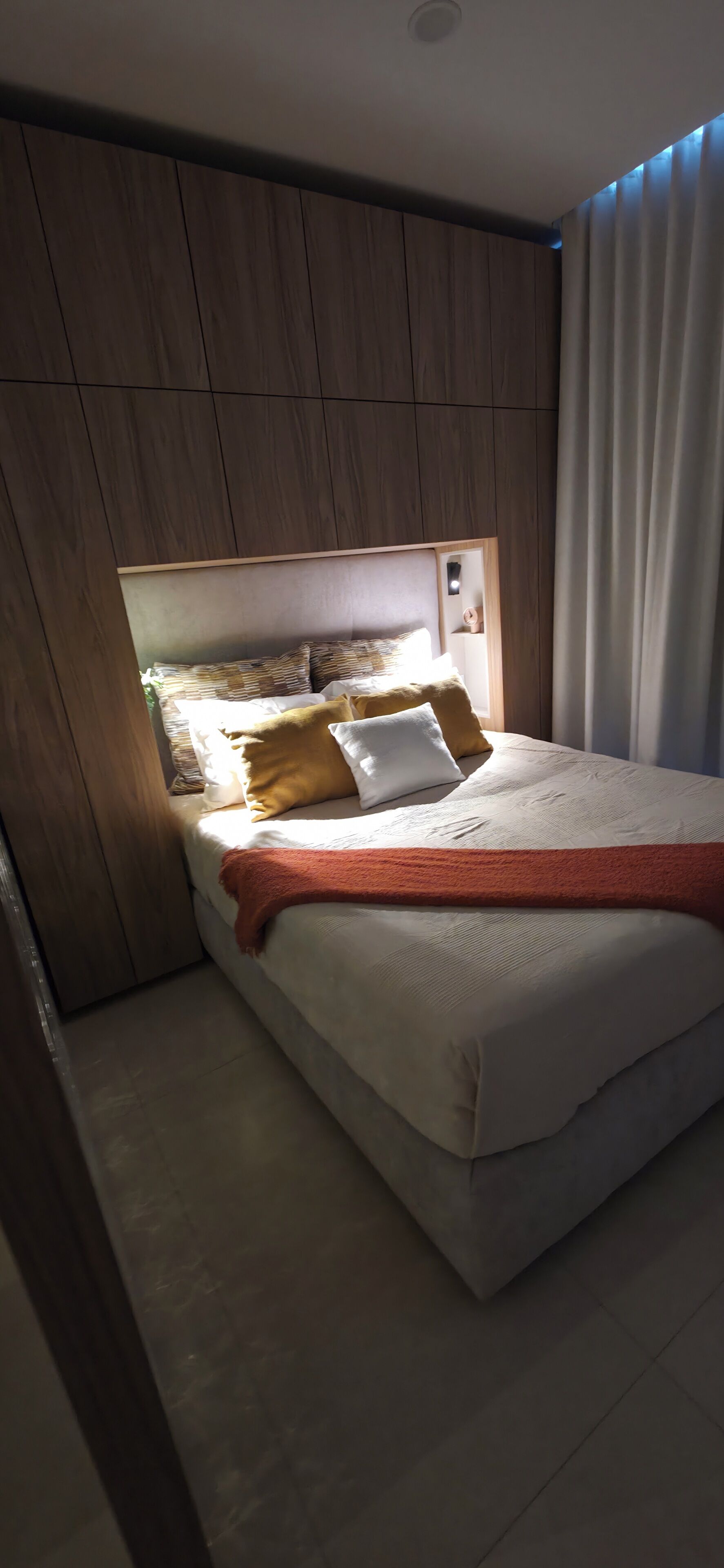 Un dormitorio con cabecera de madera, cama con colchón blanco y manta roja, y una ventana con cortinas.