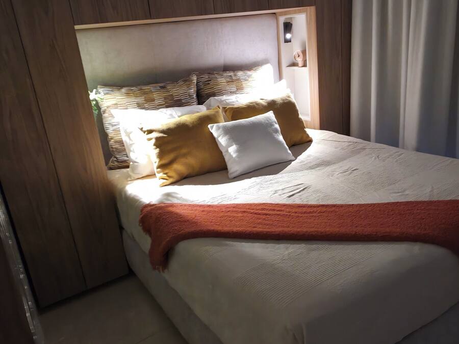 Un dormitorio con cabecera de madera, cama con colchón blanco y manta roja, y una ventana con cortinas.