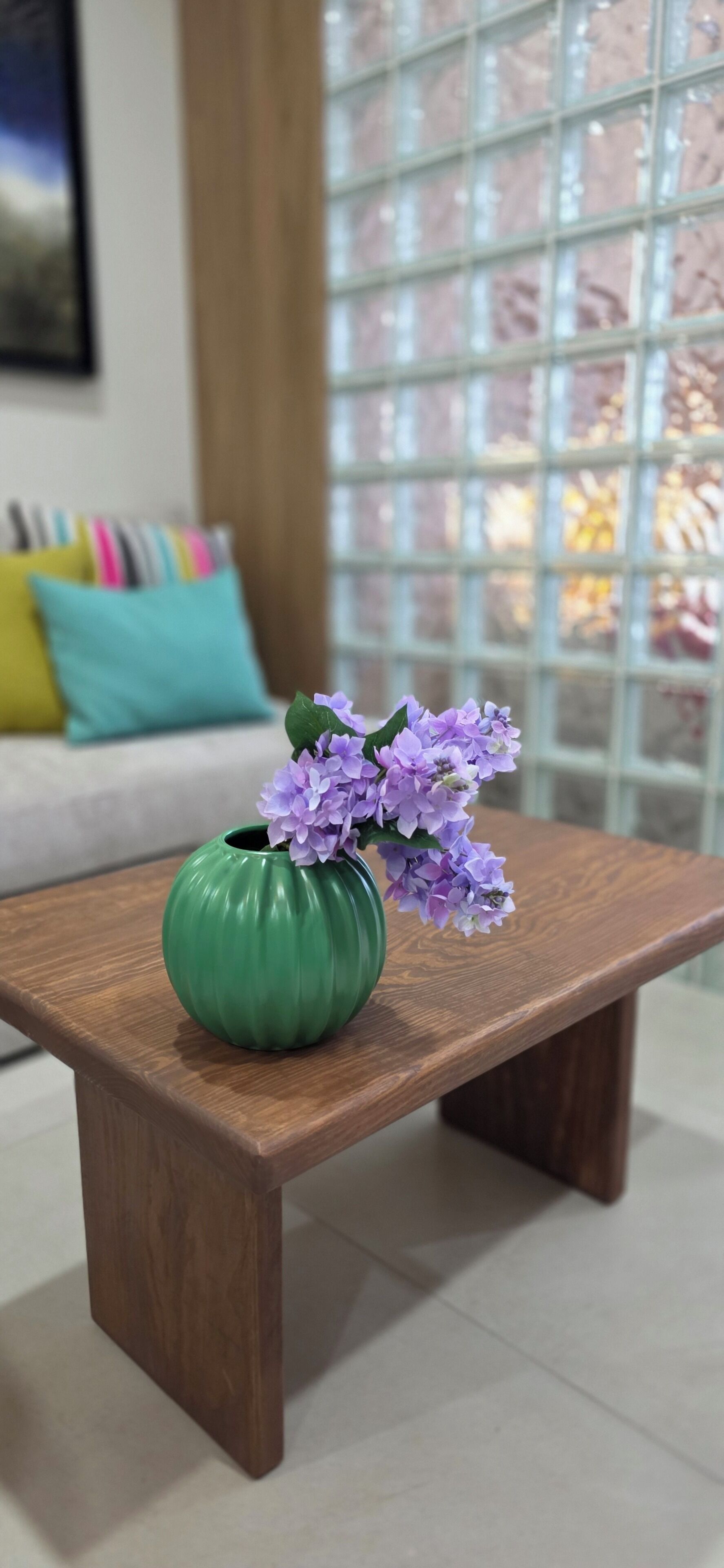 A wooden table with a green vase containing purple flowers, set against a window with a grid pattern.