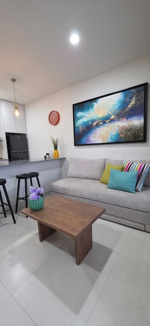 A modern living room with a sofa, a coffee table, a kitchen area, and a large abstract painting on the wall. - Ortega Living (Guadalajara)