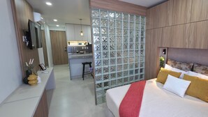 A modern hotel room with a bed, a small desk, a glass partition, and a kitchenette in the background. - Ortega Living (Guadalajara)