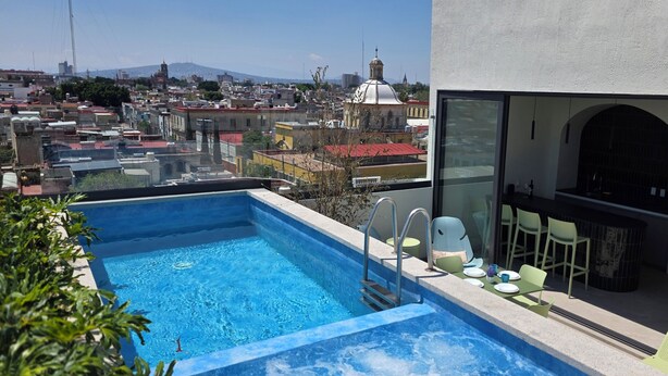 A rooftop pool with a city view, a dining area with chairs and tables, and a large arched window. - Ortega Living (Guadalajara)