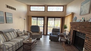 Een woonkamer met een bank, fauteuils, een salontafel en een open haard. Er zijn grote ramen en een deur die naar buiten leidt.