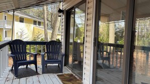 Een overdekte veranda met twee Adirondack-stoelen, een glazen deur en uitzicht op een geel gebouw op de achtergrond.