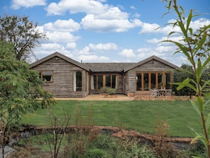 A rustic log cabin with a large glass door and windows, surrounded by a grassy yard and a small stream. - Owl Barn - Farnborough, Banbury sleeps 4 guests  in 2 bedrooms (Farnborough, Banbury)