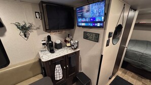 A compact living space with a kitchenette, a mounted TV displaying a streaming service, a clock, and a bed. - Cozy 1-bedroom camper in beautiful Manitowoc, walking distance to Lake Michigan (Manitowoc)
