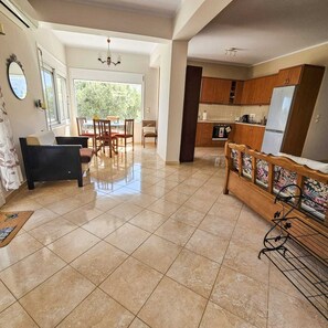 A spacious living area with a dining table and chairs, a sofa, a kitchen with wooden cabinets and a refrigerator, and a large window providing natural light. - Effie's Escape Spacious House in Zola (Argostoli)